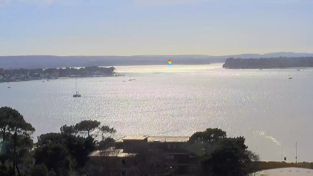 A panoramic view of a calm body of water reflecting sunlight, with small boats visible on the surface. In the background, there are hills and a shoreline lined with trees and buildings. The sky is clear, and there is a colorful shimmer of light on the water, suggesting a vibrant atmosphere.