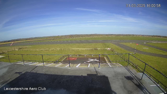 A clear blue sky is above a large, open grass field, which is part of an airfield. In the foreground, a low railing surrounds a viewing area with white marking lines on the ground. The airfield features a tarmac runway visible in the distance, with several white lines and a small red flag on the right side. The atmosphere is bright and sunny, suggesting a pleasant day. The date and time displayed indicate March 31, 2025, at 4:00 PM.