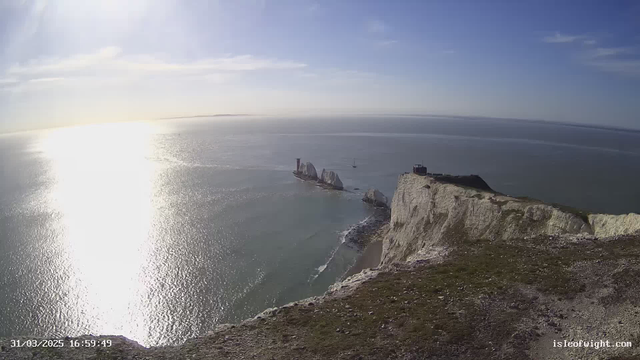A panoramic view of a coastline featuring white chalk cliffs along the shore. The sun shines brightly, creating a reflection on the calm sea. In the distance, several rock formations rise above the water, with a small sailboat nearby. The sky is clear with a few light clouds. On the cliff edge, there is a structure resembling a lookout point. The scene conveys a sense of tranquility and natural beauty.