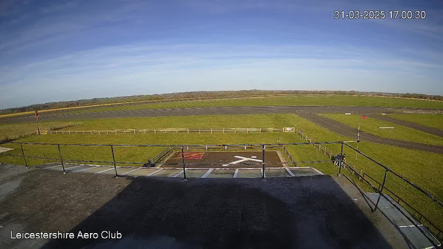 A view from a webcam at Leicestershire Aero Club, showing a wide expanse of grass-covered ground and some paved areas. In the foreground, there is a helipad marked with a large white "H" symbol and red coloring. A wooden fence borders the area. In the background, a runway is visible, leading into the distance, with green fields on either side under a clear blue sky with a few wispy clouds. The time and date displayed are March 31, 2025, at 5:00 PM.