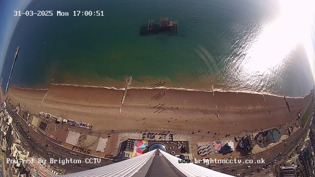 Aerial view of Brighton beach on a sunny day. The scene shows a sandy beach with a few people walking along the shore. In the water, gentle waves are visible. A pier extends into the sea from the left side of the image, while remnants of an old pier can be seen further out in the water. The sandy area is bordered by a promenade lined with beachside buildings. There are colorful structures and beach equipment visible near the shore, along with some greenery in the upper right corner. The image includes a timestamp and a logo at the bottom.