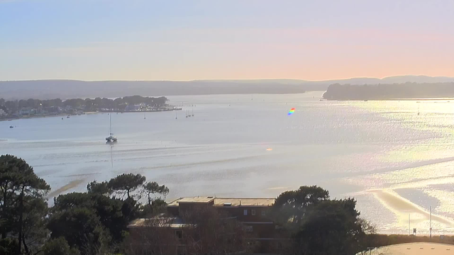 A serene waterfront scene captures a shimmering body of water under a clear sky at sunset. In the foreground, there are patches of green trees. In the middle of the water, a small sailboat glides gently, leaving a light wake behind. The horizon features distant hills and a faint outline of a coastline. Sunlight reflects off the water, creating glimmers and a soft rainbow effect in some areas. Several small boats are scattered across the water. The overall atmosphere is peaceful and idyllic.