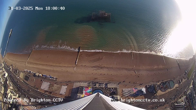 Aerial view of a beach with golden sand meeting greenish-blue water. There are waves gently lapping at the shore. Visible structures include a pier extending into the sea and beachfront buildings, some with colorful roofs. A few people are seen on the beach, and shadows are cast by the pier and structures. The date and time are displayed at the top of the image.