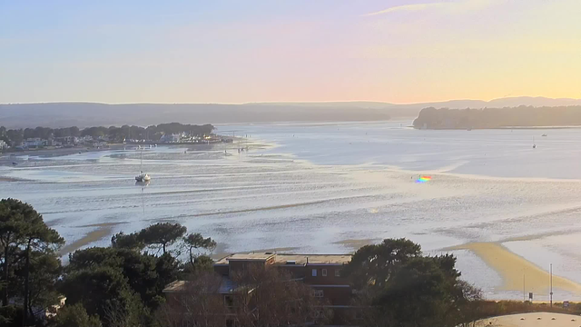 A scenic view of a calm waterway at sunset, with gentle ripples on the water's surface reflecting soft hues of orange and blue. In the distance, a small sailing boat glides peacefully through the water. Several boats are anchored along the shoreline, which is lined with trees and a few quaint buildings. The horizon showcases rolling hills under a clear sky, creating a tranquil atmosphere.