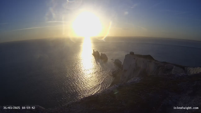 A scenic view of a coastline at sunset, featuring a vast ocean reflecting golden sunlight. In the foreground, there are rocky cliffs in shades of white and gray, and towards the center, a prominent rock formation juts out into the water. The sky displays soft blue hues with wispy clouds, while the sun radiates bright light, creating a shimmering effect on the water's surface. The date and time are displayed at the bottom of the image.