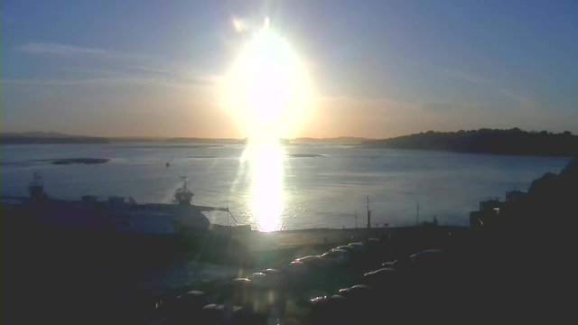 A sunny scene by the water at sunset. The sun is bright and glowing, casting a reflection on the surface of the water. In the foreground, there is a large boat docked, and several vehicles are parked along the shore. The horizon features distant hills and a clear blue sky transitioning into warmer colors as the sun sets.