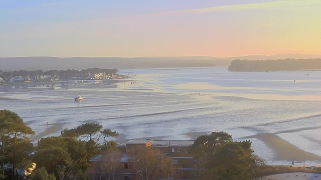 A serene coastal view at sunrise, featuring a calm waterway with gentle ripples reflecting soft hues of orange, pink, and blue. In the background, a small marina is visible with boats docked, while a few distant hills line the horizon. In the foreground, low trees and some buildings can be seen, adding depth to the scene.