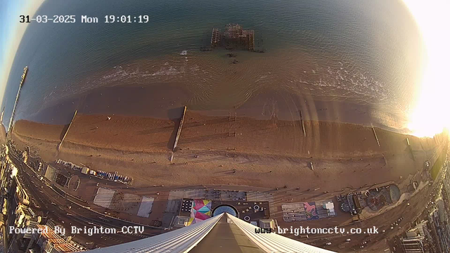 A high-angle view of a beach and shoreline during sunset. The scene includes a sandy beach extending into the water, with gentle waves lapping at the shore. Various wooden structures, possibly piers or walkways, are visible along the beach. In the foreground, there are colorful patterns on the ground, likely a public area or attraction, and some vehicles are seen parked nearby. The image captures the tranquility of the seaside with a warm golden glow from the setting sun in the background.