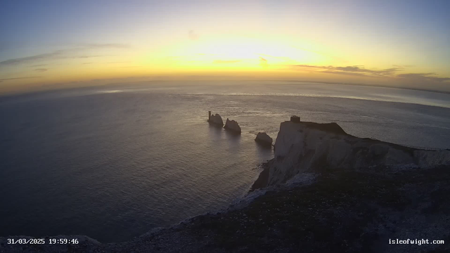 A panoramic view of a coastline during sunset. The sky transitions from soft yellow at the horizon to pale blue above, with wispy clouds scattered throughout. Below, the calm sea reflects the colors of the sky, creating a gradient effect on the water's surface. Prominent rock formations rise from the water, with one featuring a tower-like structure. The cliff to the right is steep and partially covered in vegetation, leading to a flat area with a building at the edge. The scene exudes a tranquil atmosphere, highlighting the beauty of nature.