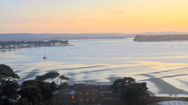 A tranquil scene of a bay during sunset. The sky is painted in soft hues of orange and pink, gently fading into a light blue at the horizon. The water reflects these colors and has gentle ripples, with a few small boats scattered across the calm surface. In the foreground, there are silhouettes of trees, and slightly to the right, a low building is visible with one window illuminated. In the distance, a line of hills frames the background, completing the serene landscape.