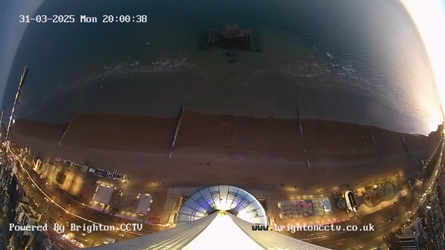Aerial view of a beach at dusk, with a dark blue ocean and shoreline visible. The beach has a sandy area with some structures, possibly beach huts or chairs, along the edge. A pier extends into the water, partially submerged. The image shows a lit promenade with small lights illuminating the walkway. The sky transitions from bright near the horizon to darker blues above. A digital timestamp is displayed in the upper left corner, indicating the date and time.