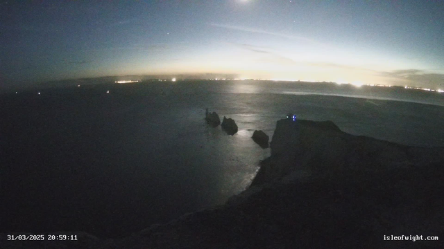A dusk scene overlooking a calm sea with large rock formations jutting out from the water on the right. The distant shoreline is illuminated with scattered lights, indicating nearby towns or structures. The sky transitions from dark to lighter shades near the horizon, where faint clouds can be seen. A slight reflection of the moonlight shimmers on the water's surface, creating a tranquil atmosphere.
