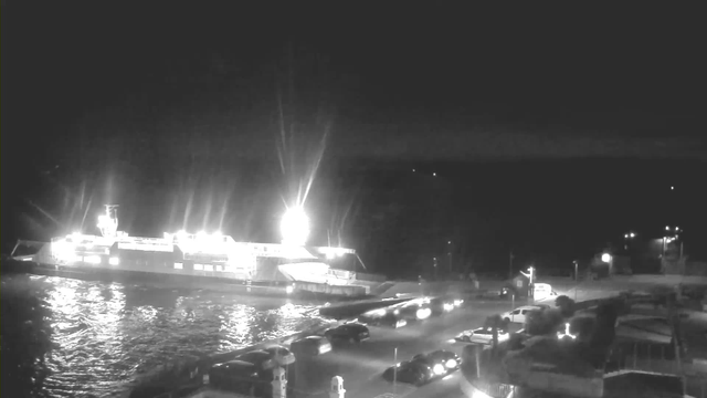 A brightly lit ferry is docked at a harbor at night, reflecting light on the water. Multiple vehicles are parked along the waterfront, and some small buildings and lights are visible in the background. The scene has a calm, maritime atmosphere, set against the dark sky.
