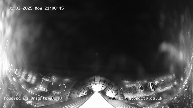 A dark, nighttime urban scene captured by a webcam. The image has a timestamp indicating it is March 31, 2025, at 21:00:45. The lower part of the image shows a wide, arched structure, possibly a building or roof, with faint light coming from an opening in the center. Surrounding the structure are blurred outlines of streets and buildings, with scattered lights indicating parked vehicles or streetlamps. The image is predominantly black and gray, creating a moody atmosphere.