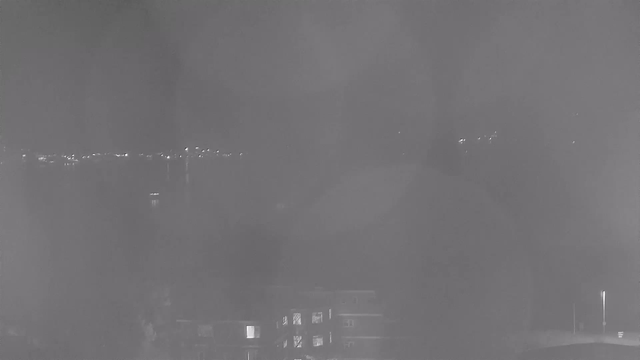 A dimly lit nighttime scene featuring a body of water reflecting faint lights from a shoreline. There are several small light sources suggesting buildings or vehicles in the distance. The foreground is less defined, with some indistinct shapes and a potential structure partially visible. Overall, the image has a grayish hue with low contrast.