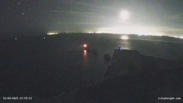 A nighttime view of a coastal scene with a body of water reflecting faint lights from a distant shoreline. The moon shines brightly in the sky, and various stars are visible. In the foreground, there is a rocky shore with a structure illuminated by blue lights. A small boat with a red light is also present in the water, near the shoreline. The overall scene is dark with limited visibility, highlighting the contrast between the light sources and the night sky.