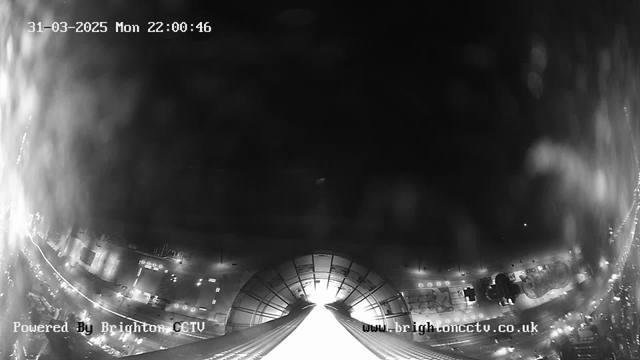 A black and white image from a webcam showing a view of a cityscape at night. The lower portion of the image features a bright light leading into the distance, possibly representing a street or pathway. Surrounding lights are visible, indicating buildings and structures, while the upper part remains mostly dark with minimal details. Text in the image shows the date and time in the top-left corner, and a watermark in the bottom-left corner indicates it is powered by Brighton CCTV.