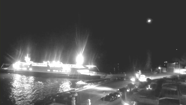 A brightly lit ferry docked at a terminal at night, with its lights reflecting on the water. Nearby, a paved area with parked cars and some structures can be seen. In the background, a few lights suggest the presence of buildings and trees along the shoreline. The sky is dark, with a faint moon visible.