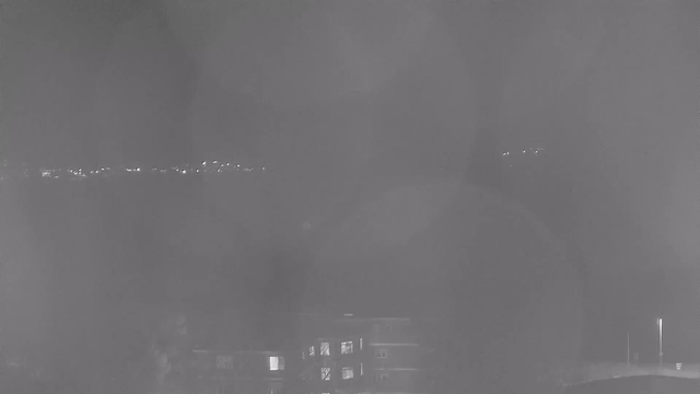 A grainy black-and-white image taken at night, showing a dimly lit scene with indistinct silhouettes of buildings in the foreground. There are faint lights along the horizon, likely from distant structures or traffic, and some blurred spots that suggest moisture on the camera lens. Overall, the image captures a dark, nighttime atmosphere with minimal visibility.