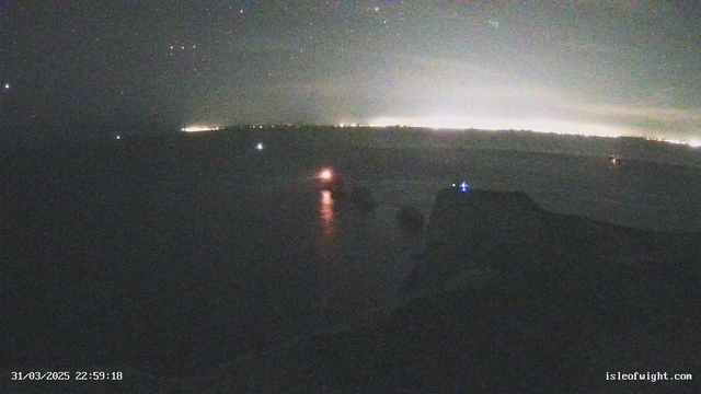 A nighttime view of a waterfront with a dark sky and faint stars. In the distance, a city skyline glows with lights on the horizon. There are multiple small lights on the water, including a brighter orange light closer to the foreground, which may indicate a boat. The shoreline is partially visible, suggesting rocky terrain. The overall scene is dimly lit with a moody atmosphere.