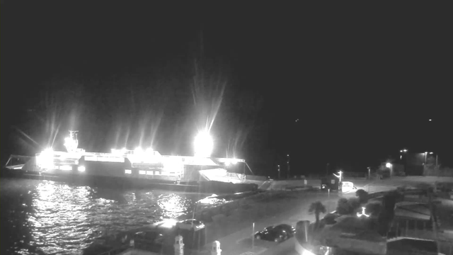 A brightly lit ferry is docked at a harbor under a night sky. The ferry is illuminated with strong lights, reflecting off the water's surface, which appears dark with ripples. To the right, there are a few parked vehicles and low structures, with additional lights illuminating the surrounding area. The scene is primarily monochromatic, emphasizing the contrast between the bright ferry and the dark surroundings.