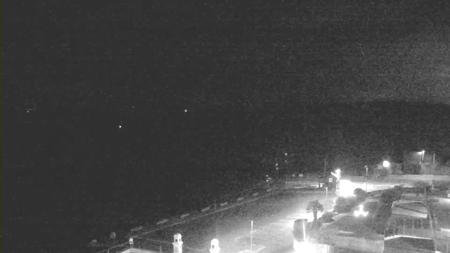 A dimly lit scene, predominantly dark with faint outlines of structures in the foreground. There are a few small, bright lights scattered throughout the image, suggesting distant objects or reflections. The background appears to be mostly empty, indicating a nighttime setting, possibly near a waterfront or coastline. There is an empty road in the lower part of the image with light sources hinting at nearby buildings or structures.