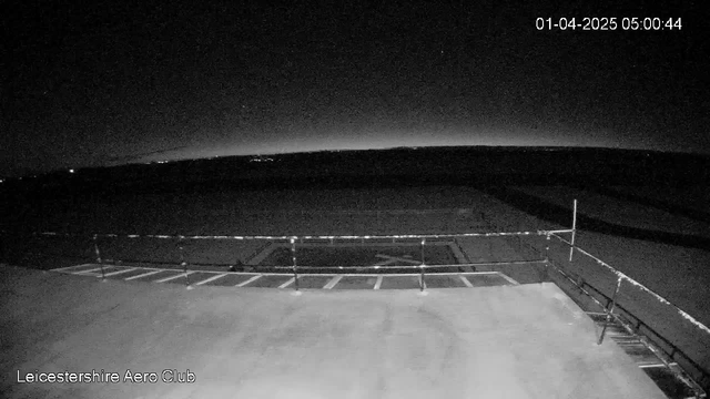 A dimly lit scene capturing a viewing platform at a small airport or aerodrome, with dark pavement visible in the foreground. A railing runs across the bottom of the image, while the horizon shows a faint glow against the dark sky, suggesting early dawn. The timestamp in the corner indicates the date and time as 01-April-2025, 05:00:44.