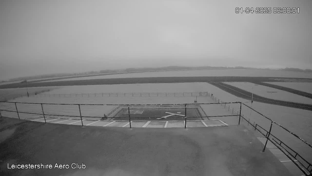 Black and white image from a webcam showing a cloudy sky and a grassy field. In the foreground, there is a fenced area with a helipad marked by an "X". The image appears foggy, limiting visibility, with flat land extending into the distance. The bottom left corner contains the label "Leicestershire Aero Club" and a timestamp indicating the date and time.