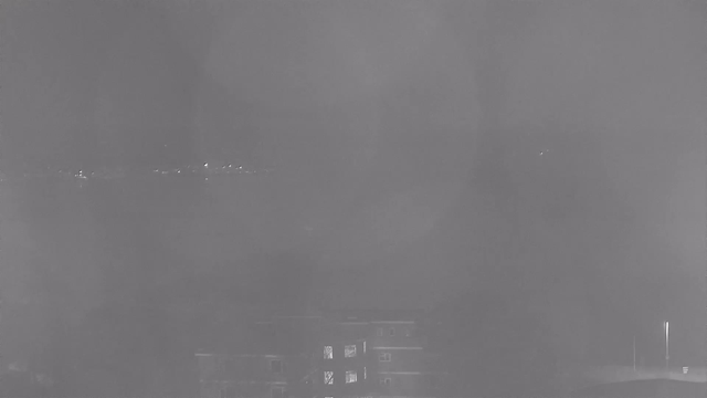 A dark, foggy scene with low visibility. In the foreground, there are outlines of buildings, with some windows faintly illuminated. In the background, a faint line of lights along a waterway suggests distant boats or a shoreline. The overall atmosphere is gray and obscured.
