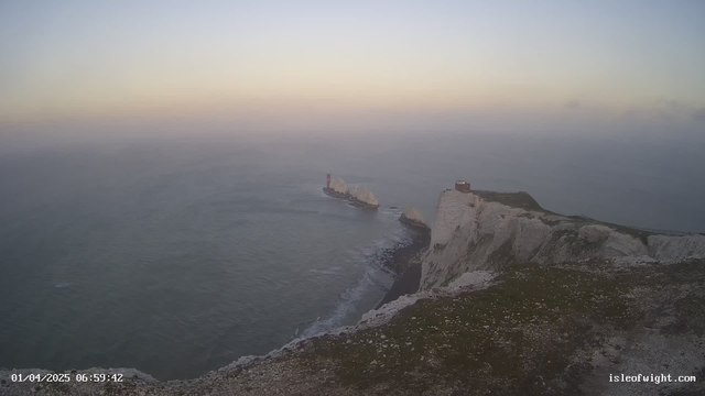 A wide view of a coastal landscape at dawn. In the foreground, there are white chalk cliffs with patches of green vegetation. In the middle distance, a small lighthouse structure can be seen on a rocky outcrop, surrounded by calm waters. The sky transitions from pale blue to soft orange as the sun rises, with a misty horizon blending into the ocean. The scene conveys a serene and peaceful atmosphere.