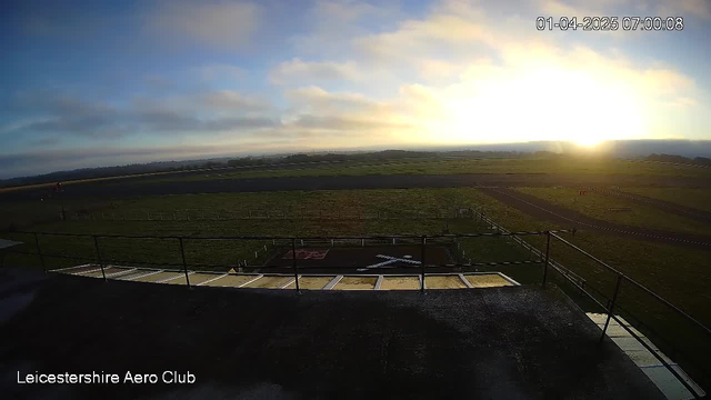 A panoramic view from a high vantage point of an airfield at sunrise. The sky is a mix of soft blue and hints of orange as the sun rises, illuminating the horizon. Below, the airfield features a grassy landscape with a runway and a marked area, including a white "X" painted on the ground, indicating a landing zone. There are wooden fences lining the airfield, with a few scattered objects, possibly aircraft tarmac. The atmosphere is calm and serene, typical of early morning.