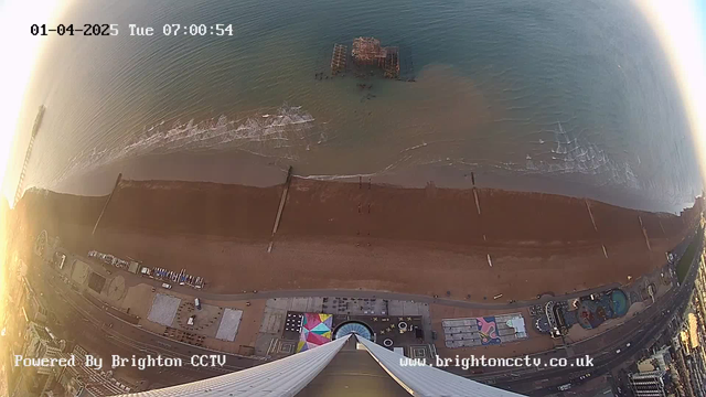 The image shows a coastal view from a high vantage point, featuring a sandy beach with scattered wooden posts and a distant pier extending into the ocean. Waves gently lap at the shore in the foreground. In the foreground, on the beach, there are several colorful beach huts and amenities. The scene is bathed in soft morning light, creating a warm atmosphere. The date and time are displayed at the top, indicating it is early morning on January 4, 2025. The bottom corner has a watermark stating it is powered by Brighton CCTV.