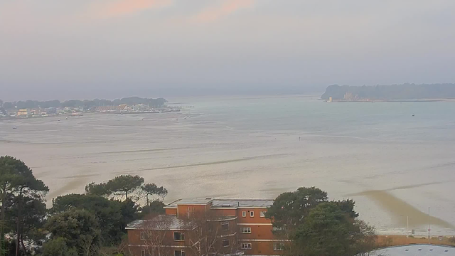 A coastal view shows a calm sea with gentle waves and boats in the water. The shoreline is lined with trees and buildings, including a few colorful structures near the beach. In the distance, a hazy outline of land can be seen, leading to a larger landmass with trees and possibly a structure on top. The sky is soft with pastel colors from the early morning light, creating a peaceful atmosphere.