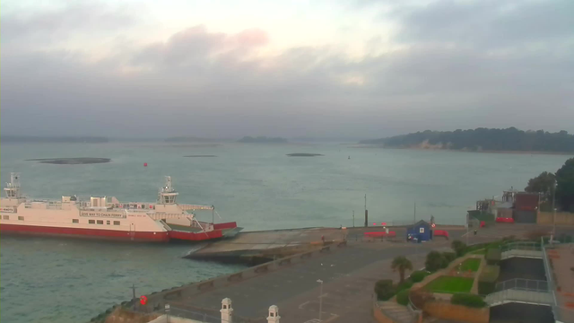 A ferry boat with a white and red color scheme is docked at a pier. The water appears calm and is a blue-gray color with a few dark patches possibly indicating submerged rocks or sandbanks. In the background, there are hazy hills and lush green trees. The sky is overcast with soft clouds, and a hint of early morning light is visible. There are red and blue structures on the dock, along with features like palm trees and landscaped areas near the water’s edge.