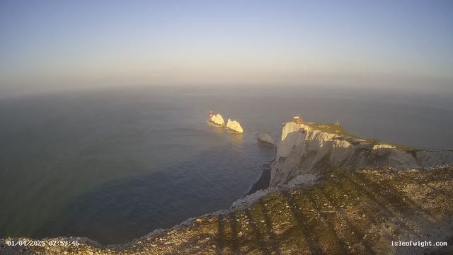 A panoramic view of a coastal landscape featuring rugged white cliffs along the sea. In the background, the sky transitions from blue to pale yellow, indicating early morning light. Two white rock formations are visible in the water, with a red lighthouse on the leftmost rock. The right side of the image shows a structure atop the cliffs, possibly a building, with greenery around it. The foreground contains rocky terrain and shadows cast by the cliffs. The water appears calm and reflects the light from the sky.