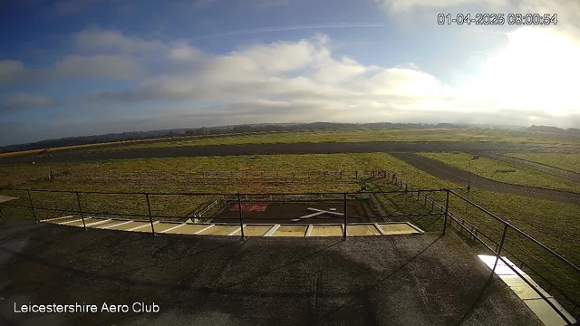 A view from a webcam at Leicestershire Aero Club shows a wide, open landscape with a grassy area in the foreground and a tarmac runway in the background. The sky is partly cloudy with a bright area indicating sunlight coming from the right side. A few fences are visible along the edges of the runway. The scene is serene and shows no aircraft or people present. The timestamp at the top indicates the date as April 1, 2025, at 08:00:54.