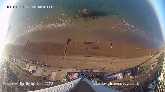 A panoramic view of a beach taken from a high elevation. The sandy shoreline stretches across the bottom of the image, showing wooden groynes protruding into the water. Gentle waves can be seen lapping at the beach. In the distance, a pier is partially submerged, with remnants visible. The water is deep blue, contrasting with the light brown sand. The image also features several colorful beach huts and structures along the coast, with the date and time displayed in the top left corner.