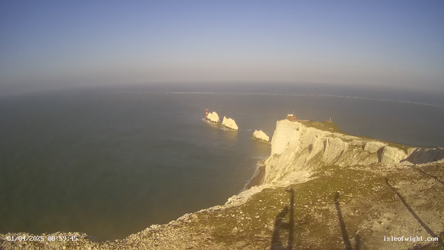 A coastal landscape is depicted with cliffs on the right side, showcasing steep white chalk formations. In the background, the sea stretches out under a clear blue sky. Two small, red lighthouses are situated on rocky outcrops, while a few boats can be seen in the water. The foreground is grassy with a visible shadow from a camera or structure at the edge of the frame.