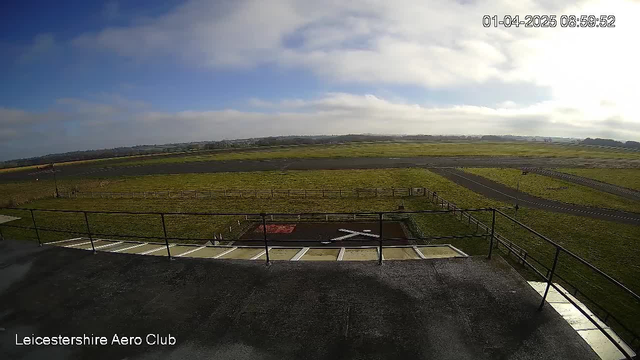 A view from the rooftop of the Leicestershire Aero Club, showing a large grassy area and an airstrip in the distance. The sky is partly cloudy with blue and gray tones. A helipad is visible in the foreground, marked with a red and white pattern. A wooden fence lines part of the grassy area, and the outskirts of the land stretch into the horizon. The scene is serene, suggesting an early morning setting.