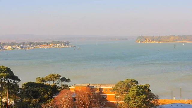 A panoramic view of a calm body of water with the horizon in the background. On the left side, there is a small coastal village with buildings and moored boats. In the distance, sailboats are scattered across the water. To the right, a small island with trees and a structure is visible. The sky is pale blue and the scene has a soft, serene appearance, suggestive of a sunny day. In the foreground, there are trees and a brick building partially visible at the bottom of the image.