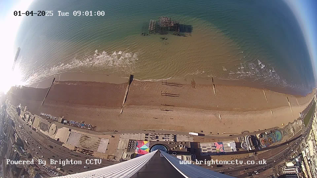Aerial view of a beach with golden sand and gently lapping waves. The water is a gradient of blue and green, reflecting sunlight. In the center, a dilapidated pier structure is partially submerged. The beach has various structures and umbrellas in a colorful pattern along the shoreline. The image is taken from above, looking down toward the coast, with a portion of a modern building visible in the foreground. The time is displayed as 09:01 on April 1, 2025, and a watermark indicates it is powered by Brighton CCTV.