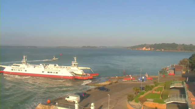 A white and red ferry is approaching a dock on a calm body of water, under a clear blue sky. In the background, a small boat can be seen moving across the water. To the right, there is a landscaped area with grassy patches and a few buildings, while the shore features a green tree line.