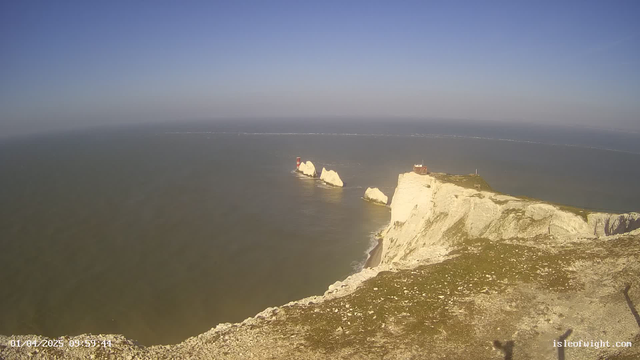 A coastal view featuring white chalk cliffs extending into the ocean. In the water, there are two prominent rock formations with red and white striped lighthouses. The sky is clear with a bright blue hue, and a few boats are visible on the horizon, creating white foam trails as they move. The foreground shows rocky terrain with sparse vegetation.