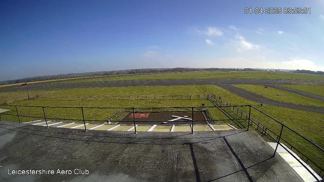 A clear blue sky stretches across the top of the image, with a few wispy clouds. Below, an expansive green field is visible, bordered by a low wooden fence. In the foreground, there is a helipad marked with a large white cross and a red square. A bit further into the scene, an asphalt runway runs parallel to the field, leading into the distance where trees can be seen lining the horizon. The timestamp in the corner reads 09:59:51 on April 1, 2025, and there’s a label at the bottom indicating "Leicestershire Aero Club."