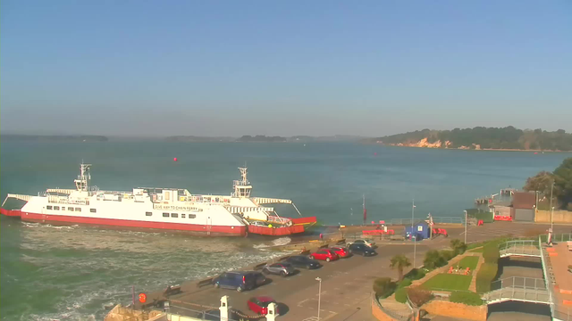A ferry is docked at a terminal on the water, with a clear blue sky in the background. The ferry is primarily white with red accents. In the foreground, several parked cars of various colors are visible on a paved area. To the right, there are green landscaped areas, and a small building can be seen next to the water. On the far side of the water, there are distant trees and cliffs.