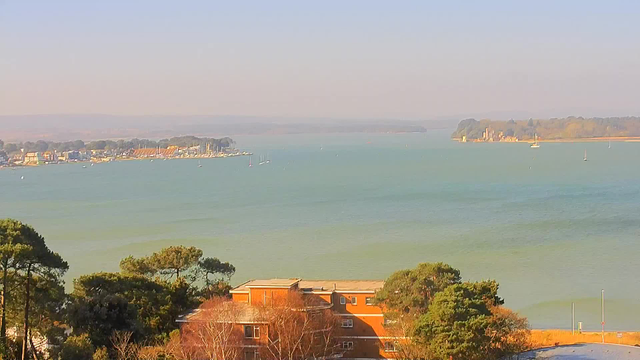 A scenic view of a calm body of water, where the ocean meets the horizon. On the left side, there are small boats and a shoreline with a row of buildings, including colorful houses and a marina. In the distance, more boats can be seen sailing. To the right, an island with trees and a structure resembling a castle is visible. The sky is clear with a soft, pale blue hue, and the light creates a warm atmosphere over the scene. In the foreground, there are trees and a brick building with a flat roof.