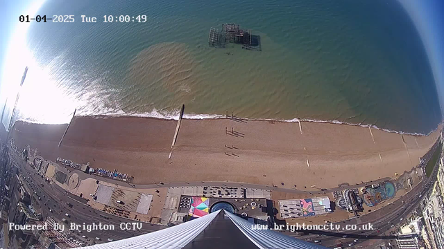 Aerial view of a beach with sandy shoreline and gentle waves, under a clear blue sky. In the foreground, there are several people walking along the beach. A pier structure is visible offshore, partially submerged in the water. The beach area features various attractions, including colorful amusement rides and marked zones. The image also has a timestamp in the upper left corner indicating the date and time.