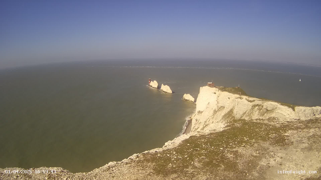 A coastal scene featuring white chalk cliffs extending into the ocean. In the distance, a group of buildings, which likely include lighthouses, is situated on rocky outcrops surrounded by water. The sky is clear with a few wispy clouds, transitioning from blue to a lighter hue near the horizon. Gentle waves can be seen lapping at the base of the cliffs. The foreground shows a grassy area leading to the cliff edge.