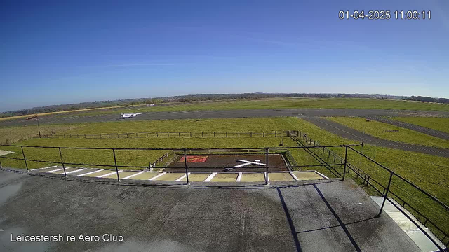 A view from a webcam at Leicestershire Aero Club shows a grassy airfield under a clear blue sky. In the foreground, there is a flat rooftop area with a railing and a helipad marked with a white cross and a red square. In the background, a small airplane is parked on the runway, which is bordered by green grass and wooden fencing. The scene conveys a tranquil, sunny day at the aviation club.