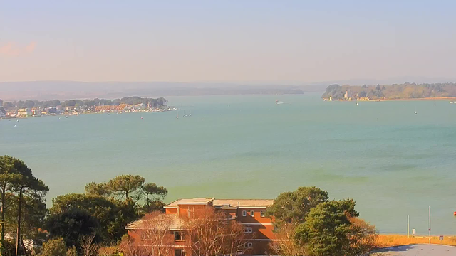A panoramic view of a calm bay, featuring turquoise blue water under a clear sky. In the background, a line of small boats is moored along the shoreline, which includes various buildings, likely homes or businesses. To the left, there are trees and greenery, and further in the distance, an island can be seen, which has a structure, possibly a castle or lighthouse, on it. The overall scene conveys a peaceful coastal atmosphere.